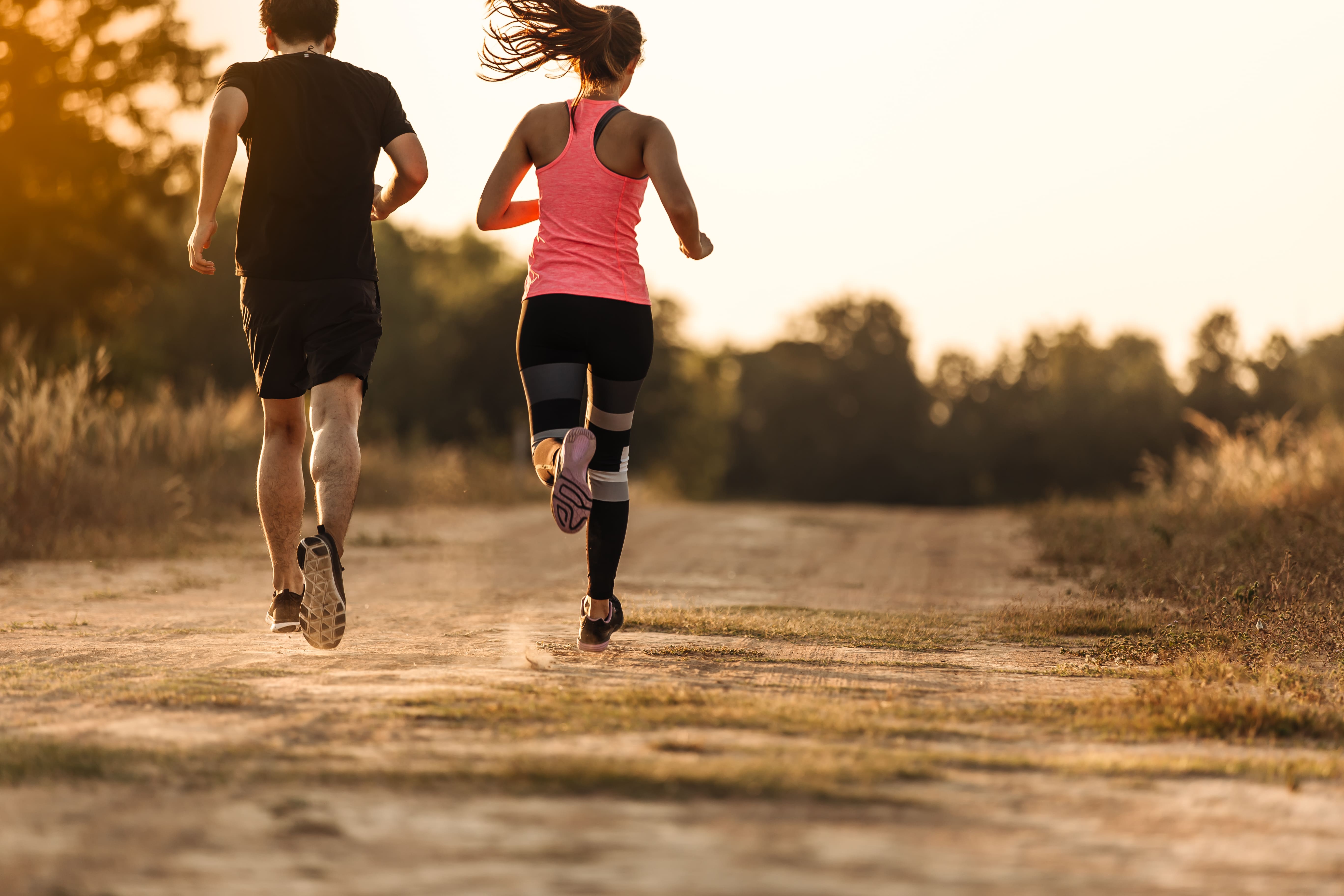 A imagem mostra um casal jovem correndo em uma trilha de terra ao ar livre durante o pôr do sol. A mulher usa uma regata rosa, leggings esportivas e tênis de corrida, enquanto o homem veste uma camiseta preta, shorts e tênis esportivos. O cenário é cercado por vegetação e árvores, com a luz dourada do sol iluminando o ambiente, criando uma atmosfera agradável e tranquila.
